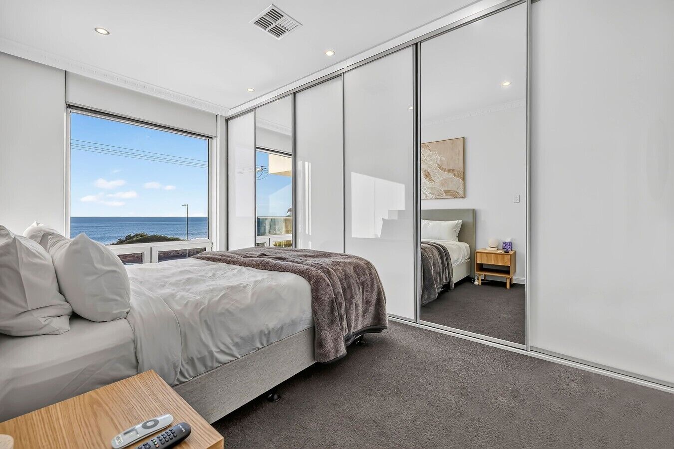 A modern bedroom with a large bed, a bedside table, a nightstand, and a view of the sea.