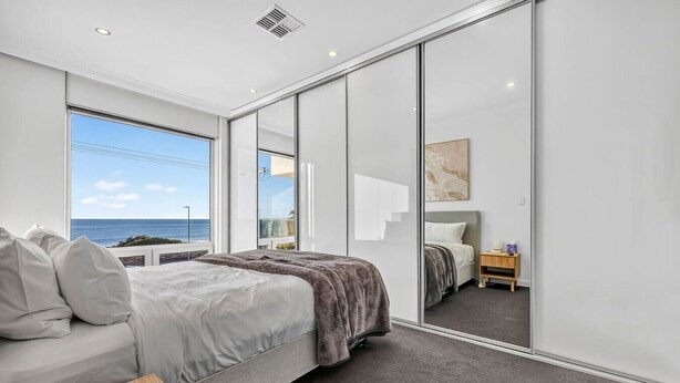 A modern bedroom with a large bed, a bedside table, a nightstand, and a view of the sea. - Sunset Sands on Esplanade | By Host Solutions (West Beach)