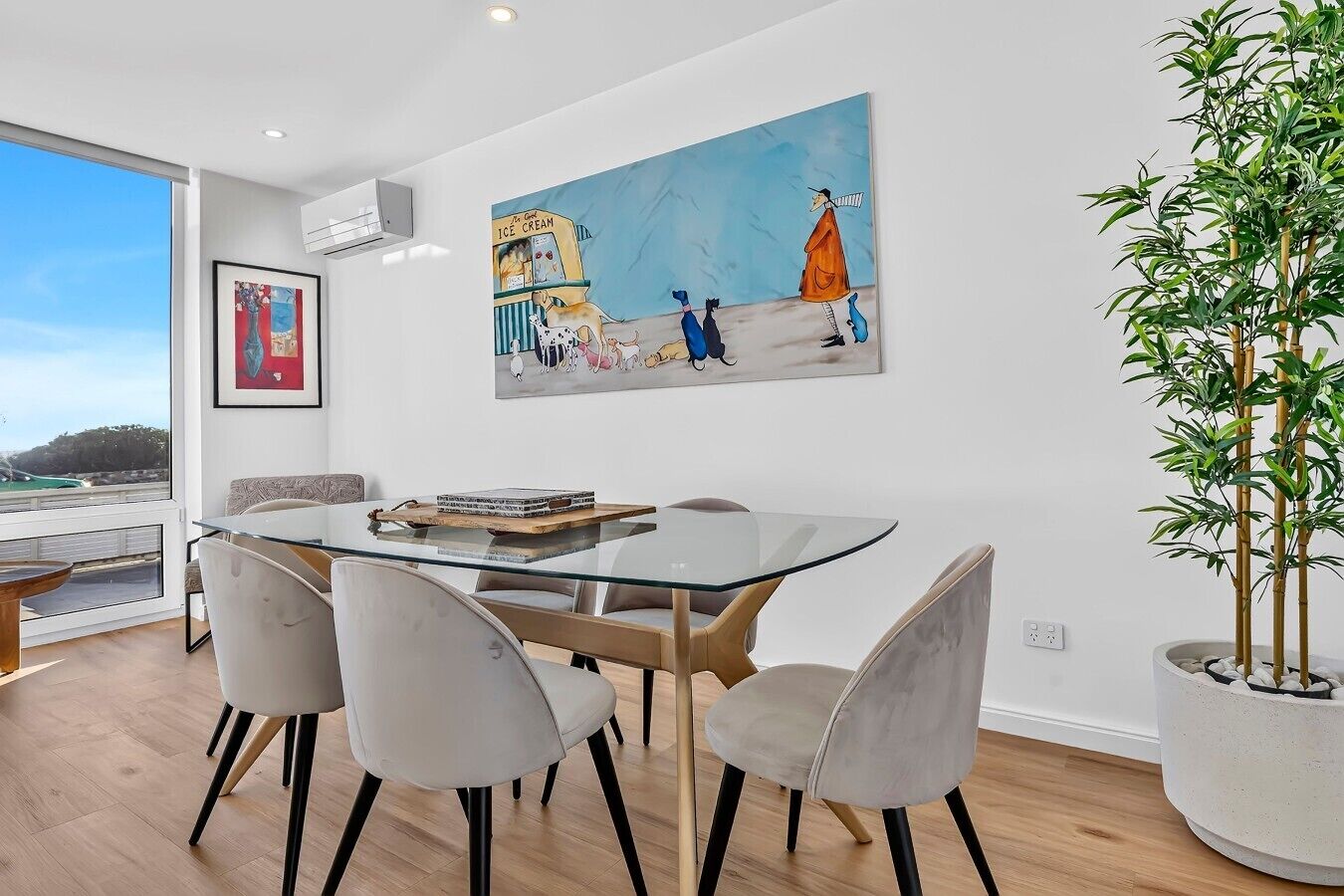 A modern dining area with a glass table, cushioned chairs, and a large painting of a whimsical scene.