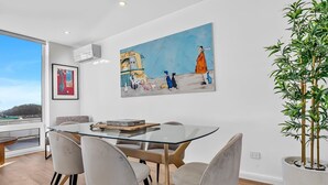 A modern dining area with a glass table, cushioned chairs, and a large painting of a whimsical scene. - Sunset Sands on Esplanade | By Host Solutions (West Beach)