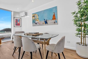 A modern dining area with a glass table, cushioned chairs, and a large painting of a whimsical scene.
