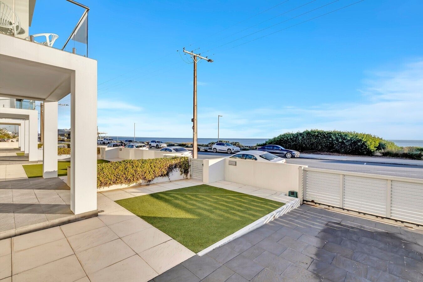 A modern building with a flat roof, a small lawn, and a view of the sea and parked cars.