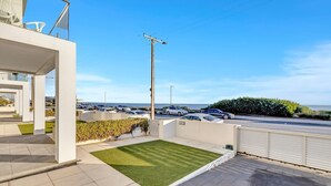 A modern building with a flat roof, a small lawn, and a view of the sea and parked cars. - Sunset Sands on Esplanade | By Host Solutions (West Beach)