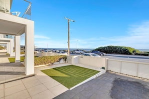 A modern building with a flat roof, a small lawn, and a view of the sea and parked cars.