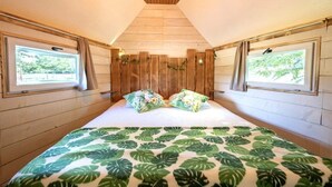 A bedroom with a wooden slat headboard, two windows with curtains, a bed with a tropical leaf-patterned bedspread, and a wooden ceiling. - ZEcamping piscine chauffée dog friendly sauna cabane BBQ clim 19m2 (Chassenard)