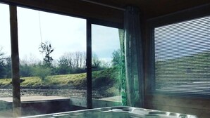 A hot tub with a view of a grassy landscape through large windows. - ZEcamping piscine chauffée dog friendly sauna cabane BBQ clim 19m2 (Chassenard)