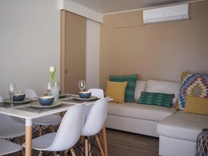 A modern living room with a dining area. There is a white sofa with colorful cushions, a dining table with chairs, and a vase with a flower. - ZEcamping 4 étoiles toboggans locatif TV LV clim 54m2 (Regencós)