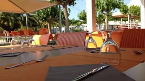 An outdoor dining area with orange chairs and tables, clear glass wine glasses, and a metal fork on a napkin.
