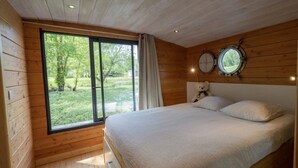A bedroom with a large window, wooden walls, a bed, a teddy bear, and a round mirror on the wall. - ZEcamping 3 étoiles piscine couverte animaux acceptés locatif clim (Chamberet)