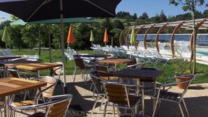 An outdoor seating area with tables and chairs, umbrellas, and a pool in the background. - ZEcamping 3 étoiles piscine couverte animaux acceptés locatif clim (Chamberet)