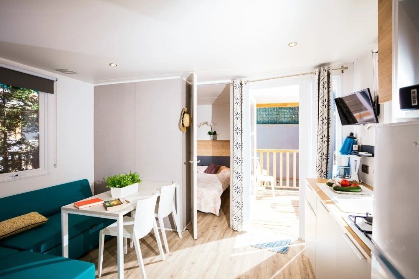 A modern, well-lit room with a teal sofa, a dining table with chairs, a kitchen area with a refrigerator, and a balcony with a view.