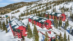 A red lodge with multiple floors and a green roof, surrounded by snow-covered trees and a mountain backdrop.