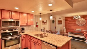 A kitchen with wooden cabinets, stainless steel appliances, granite countertops, pendant lights, and a clock on the wall.