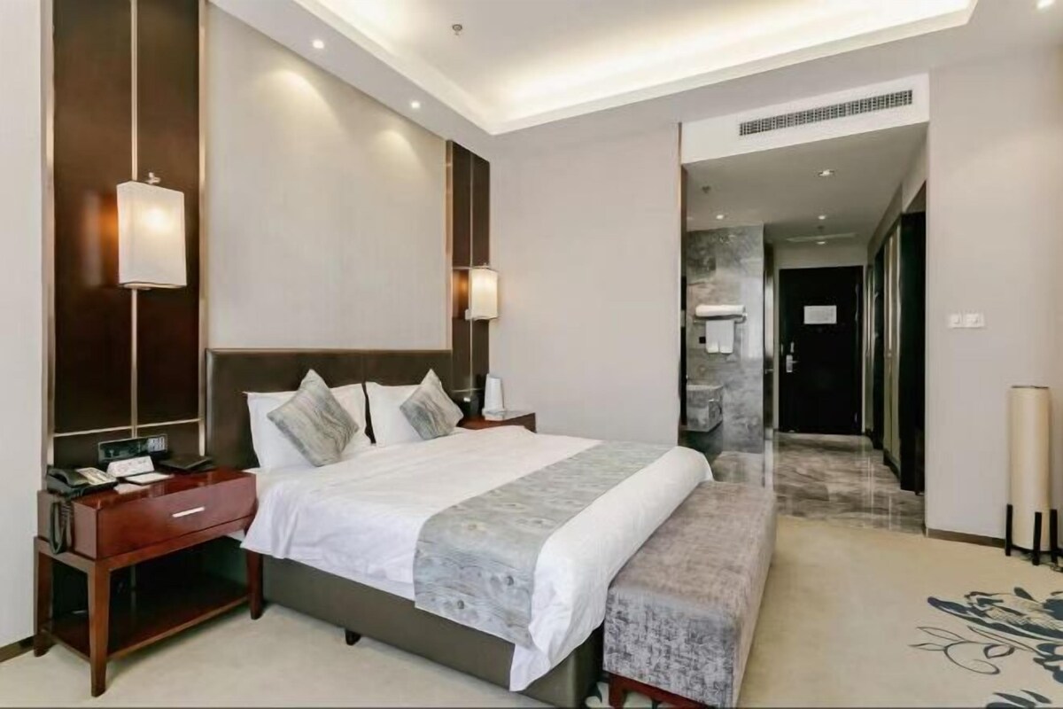 A modern hotel room with a large bed, bedside tables, a bench, and a hallway leading to a bathroom.