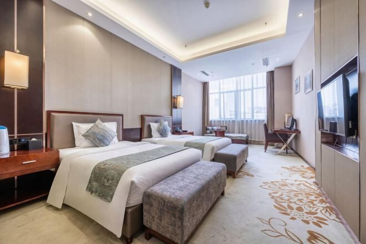 A hotel room with two beds, a desk, a TV, and a large window.