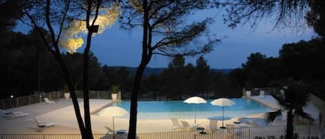 En swimmingpool med solstolar och parasoller, omgiven av träd och ett staket.