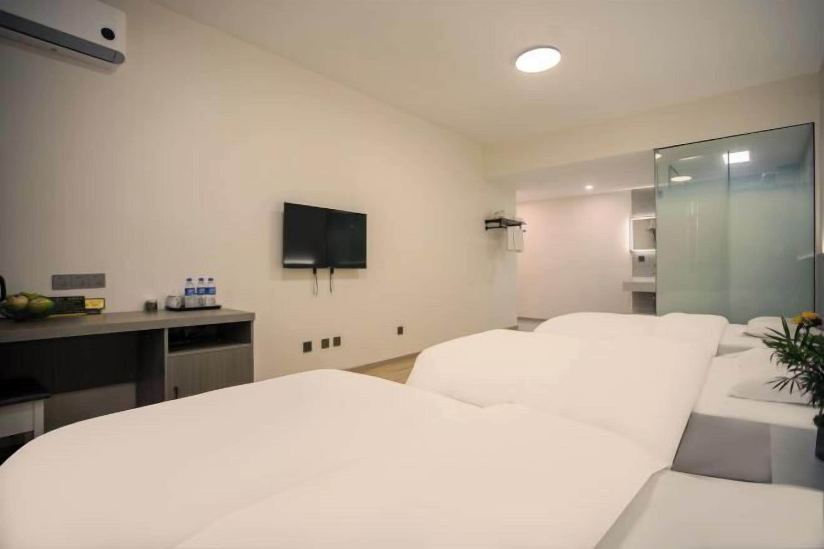 A hotel room with two beds, a flat-screen TV mounted on the wall, a small desk with a computer and a water dispenser, and a glass partition wall.