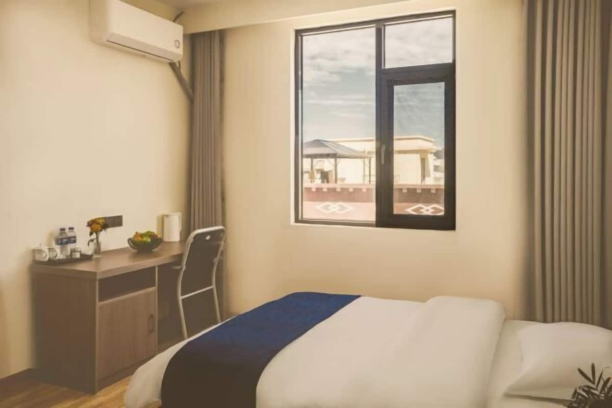 Een hotelkamer met een bed, een bureau, een airconditioner, een raam met gordijnen en uitzicht op het omliggende landschap.