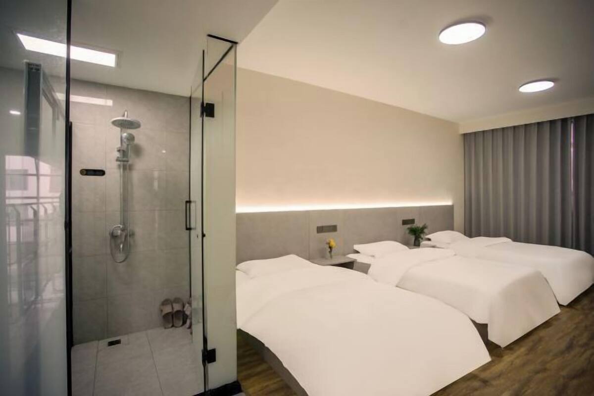 Een hotelkamer met een douchecabine van glas, twee bedden met witte lakens, een nachtkastje met een vaas en een groot raam met gordijnen.