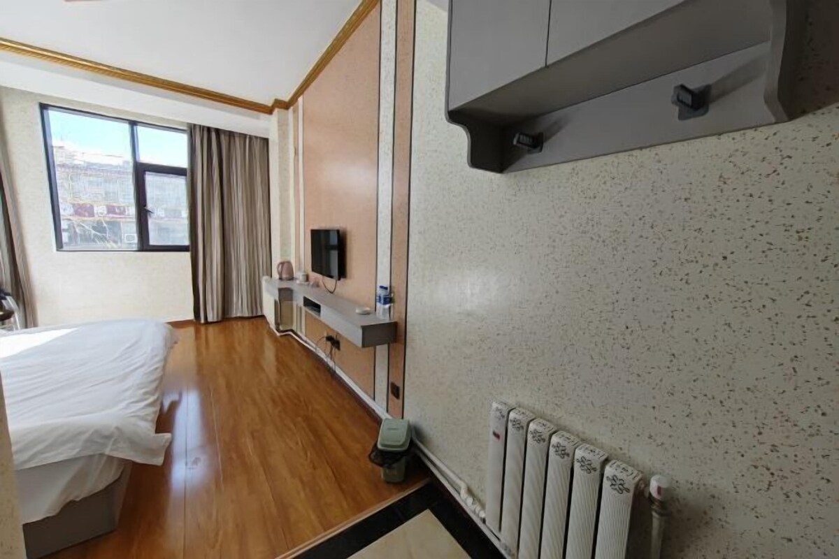A hotel room with a bed, a TV, a small table with a bottle, a radiator, and a window with curtains.