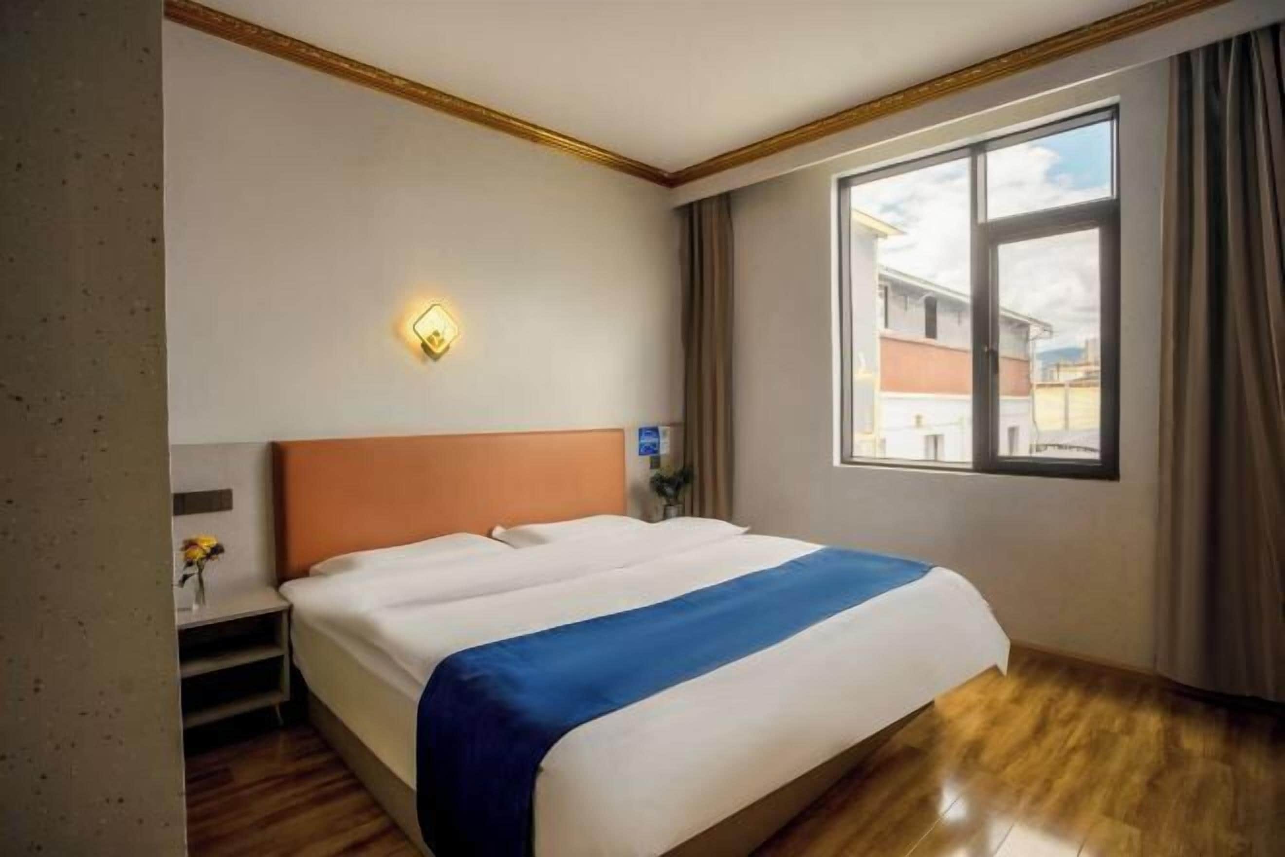 A hotel room with a large bed, a nightstand, a wall-mounted light, and a window with curtains.