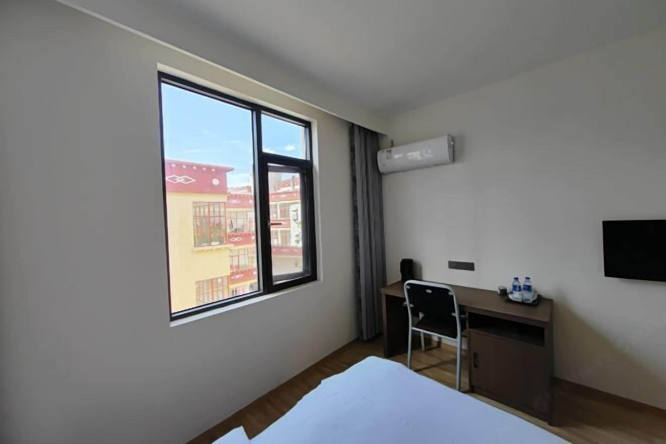 A hotel room with a large window, a desk, a chair, a bed, and an air conditioning unit.