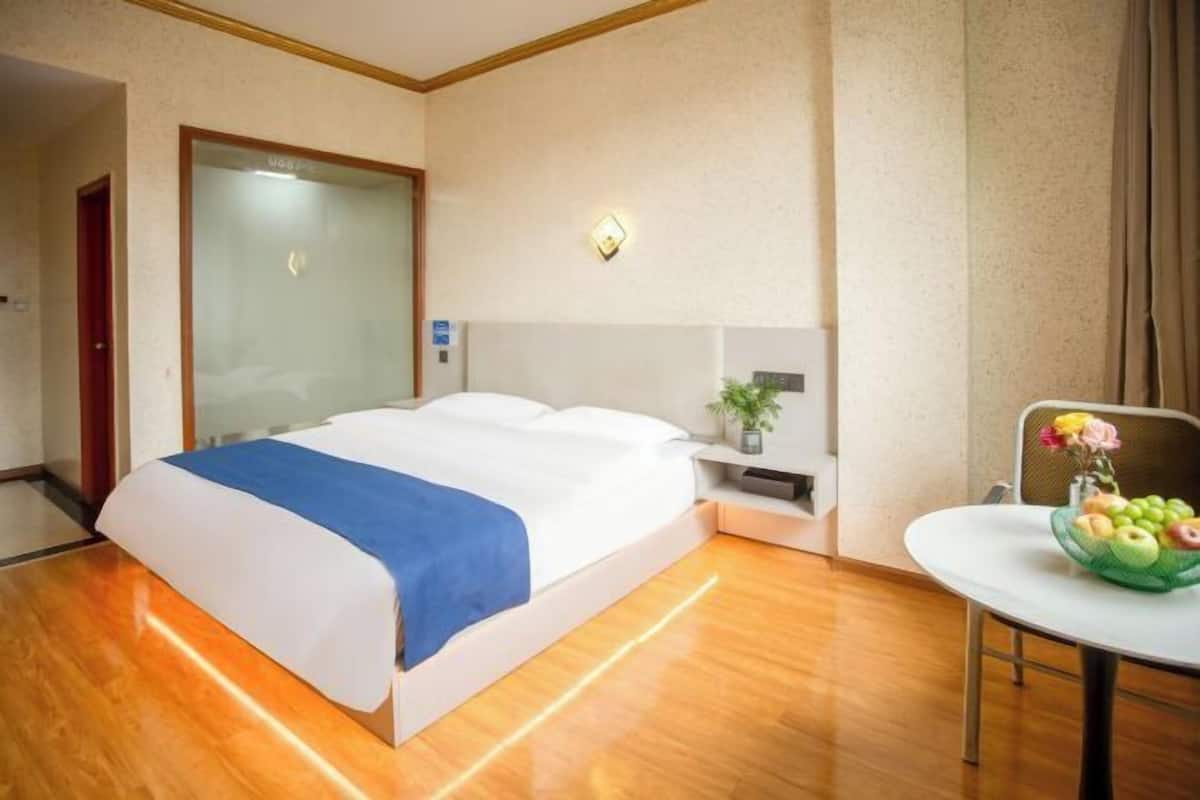 Een hotelkamer met een groot bed, een nachtkastje met een lamp, een kleine tafel met fruit, en een stoel.