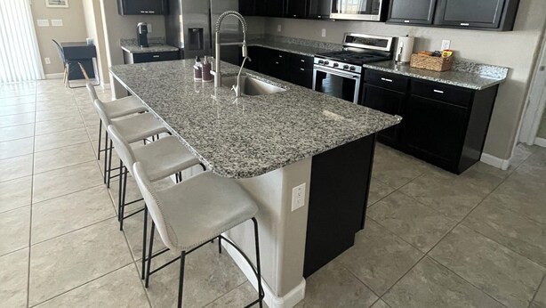 A modern kitchen with dark cabinets, a granite countertop, stainless steel appliances, and a bar stool seating area. - Spacious, Family-Friendly Home in Maricopa (Maricopa)