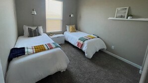 A bedroom with two beds, a window with blinds, a nightstand, a lamp, a shelf with a picture, and a carpeted floor. - Spacious, Family-Friendly Home in Maricopa (Maricopa)