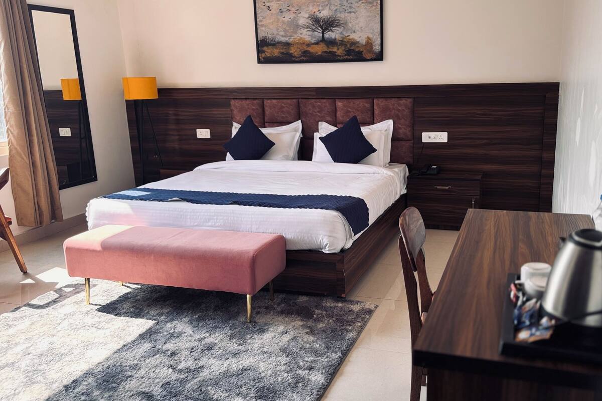 A neatly arranged hotel room with a large bed, a wooden nightstand, a desk with a kettle, and a painting on the wall.
