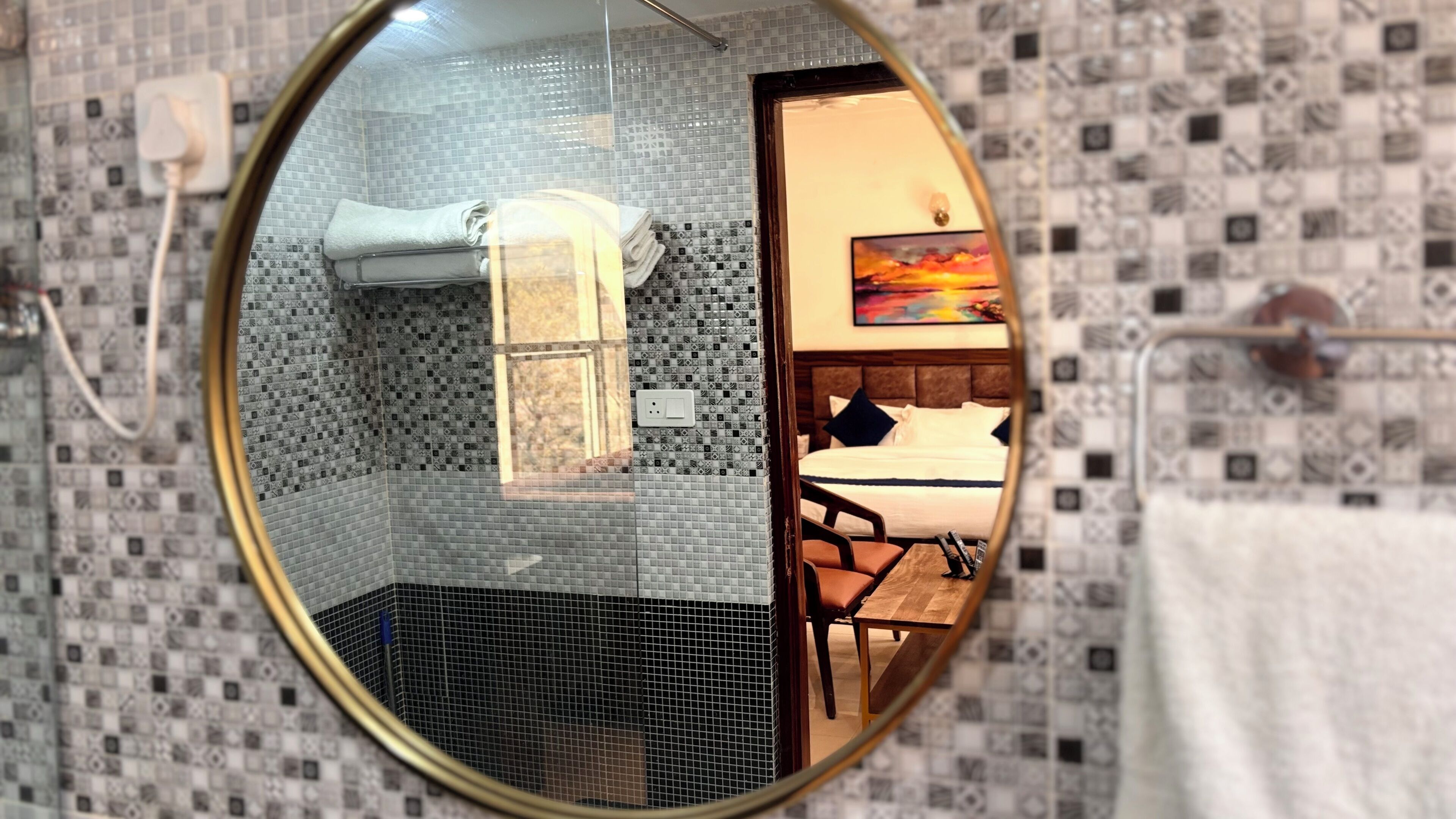 Un baño con pared de azulejos, un perchero y un espejo que refleja un dormitorio con cama, mesita de noche y un cuadro.