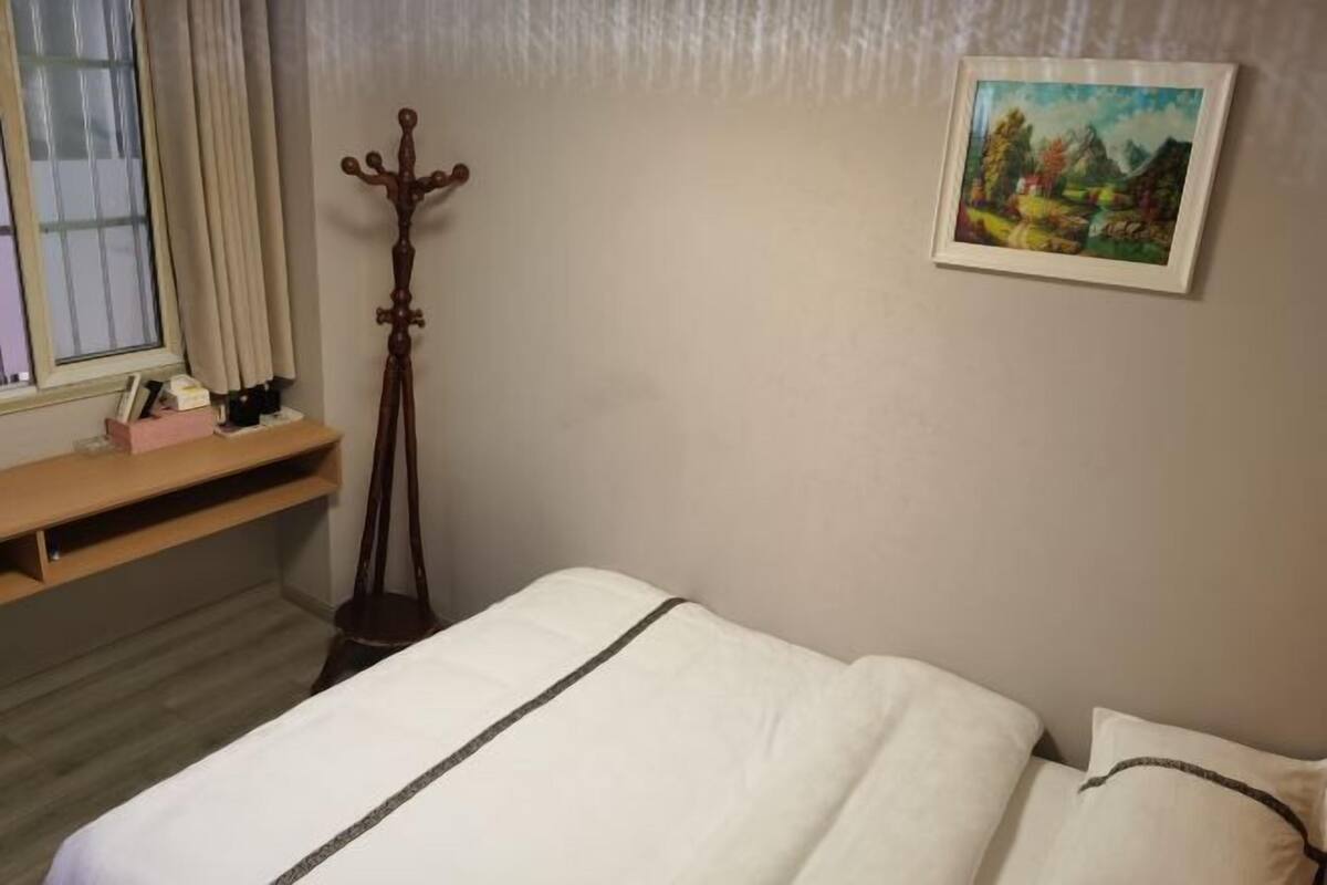 Een slaapkamer met een bed, een houten kapstok, een schilderij aan de muur en een raam met gordijnen.