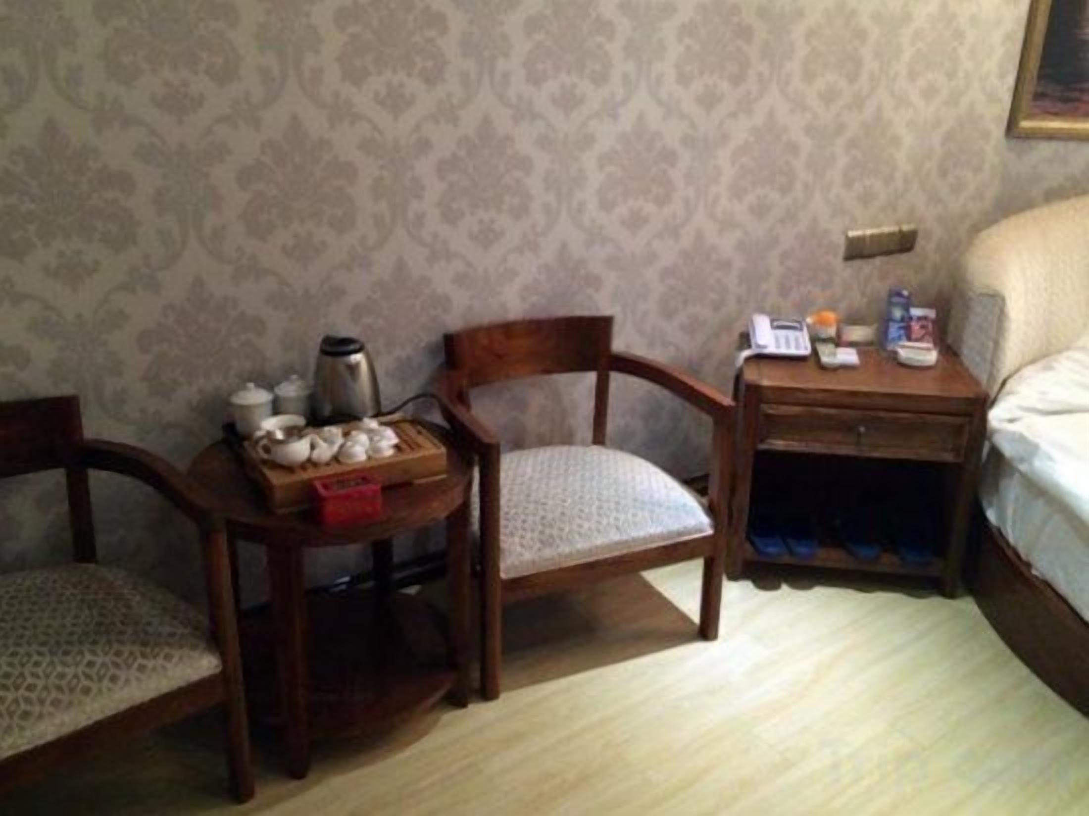 Quarto com duas cadeiras de madeira, uma mesa pequena com uma bandeja de chá e uma mesinha de cabeceira com um telefone e uma garrafa.