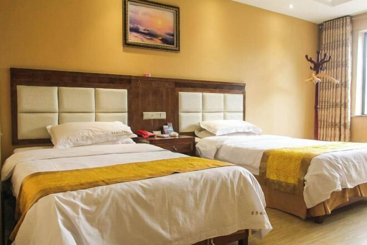 Habitación de hotel con dos camas, cabecera de madera, mesita de noche, un cuadro en la pared y una ventana con cortinas.