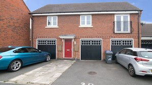 A two-story brick building with a red door, two garage doors, and a small balcony. - Lovely Two Bed detached house with Parking (Tipton)