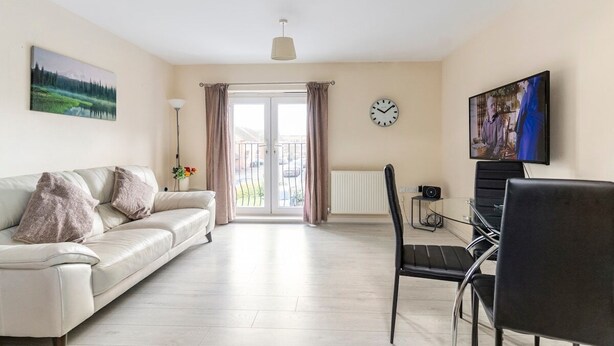 A living room with a sofa, a chair, a TV, a clock, and a window with curtains. - Lovely Two Bed detached house with Parking (Tipton)