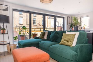 Ein modernes Wohnzimmer mit einem grünen Ecksofa, einem kleinen orangefarbenen Hocker und einer Stehlampe. Mehrere Kübelpflanzen und Blick auf ein Backsteingebäude durch große Fenster.