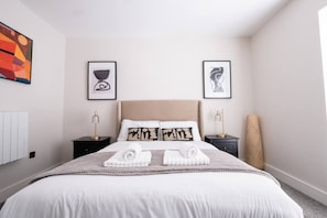 A hotel room with a large bed, two bedside tables with lamps, a radiator, and two framed artworks on the walls. - Luxury City Centre 4BR House | Sleeps 10 | Free Parking By Torr Property Group (Merseyside)