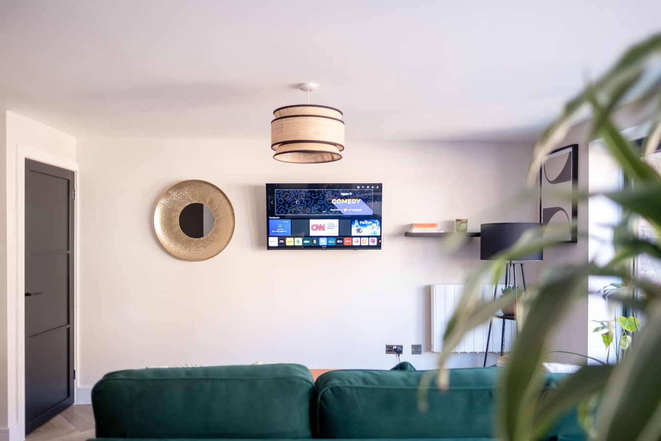 Un saló modern amb un sofà verd, un televisor penjat que mostra diversos canals, una obra decorativa a la paret i un lluminari al sostre.