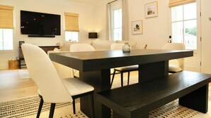 A modern dining room with a dark wooden table, white upholstered chairs, and a television mounted on the wall.