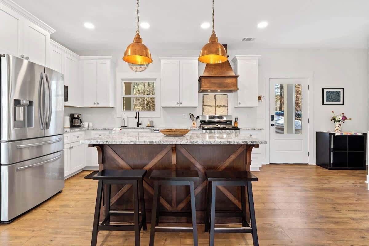 A modern kitchen with white cabinets, stainless steel appliances, a marble countertop, and dark wooden stools.