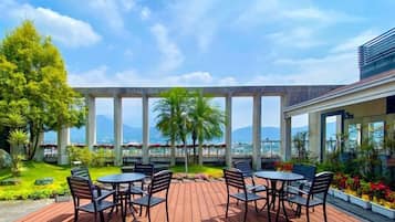 An outdoor patio with tables and chairs, surrounded by greenery and a view of the cityscape.