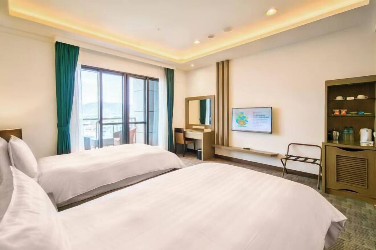 Habitación de hotel con dos camas, un escritorio, una silla, un televisor, un espejo y una ventana con cortinas.