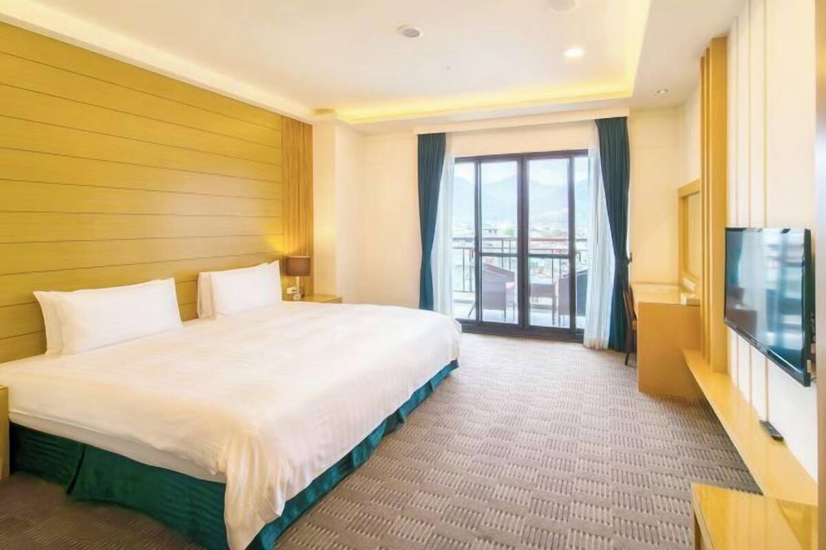 Habitación de hotel con una cama grande, televisor, balcón con vistas y ventana con cortinas.