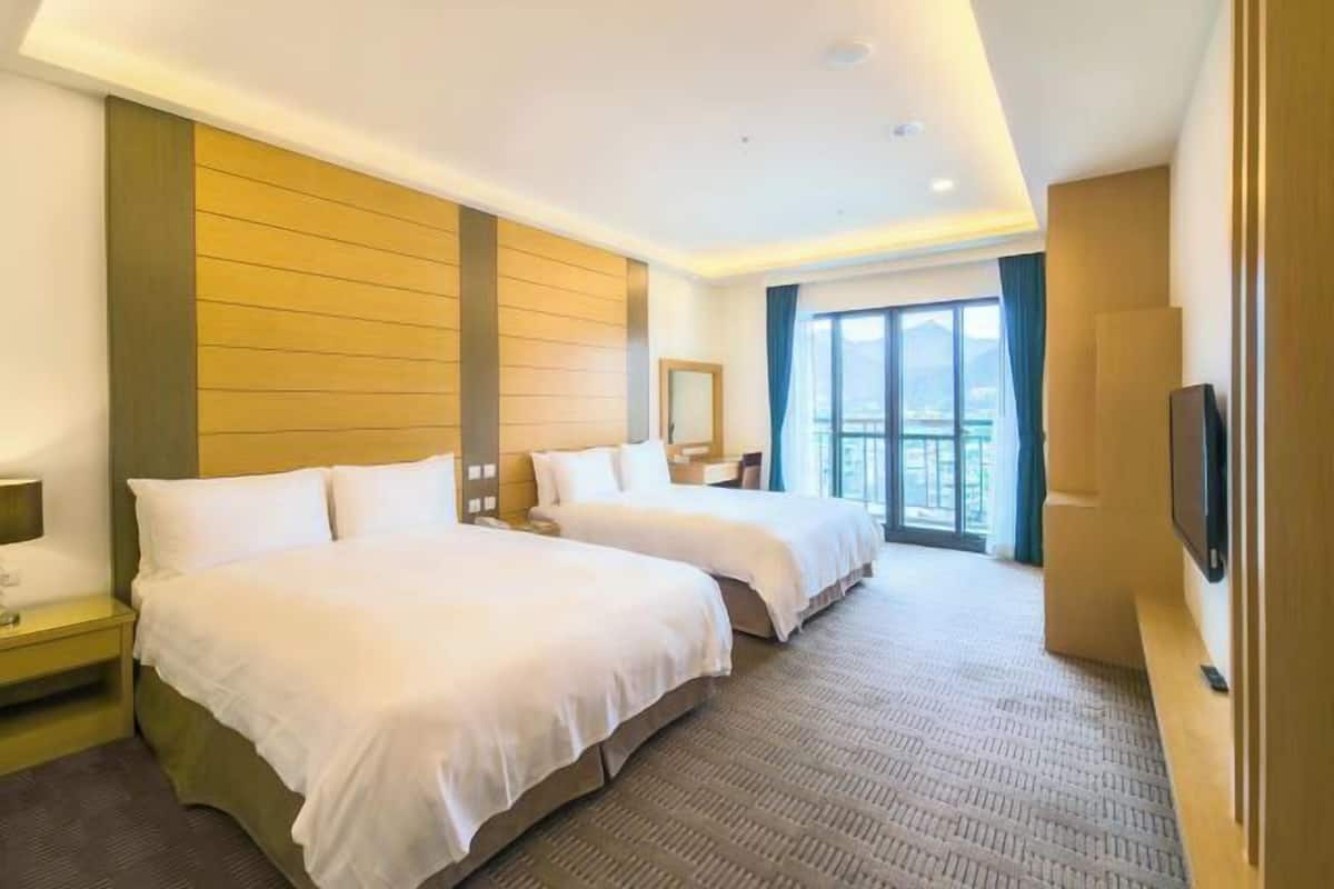 Habitación de hotel con dos camas, televisión, ventana grande y vista a la ciudad.