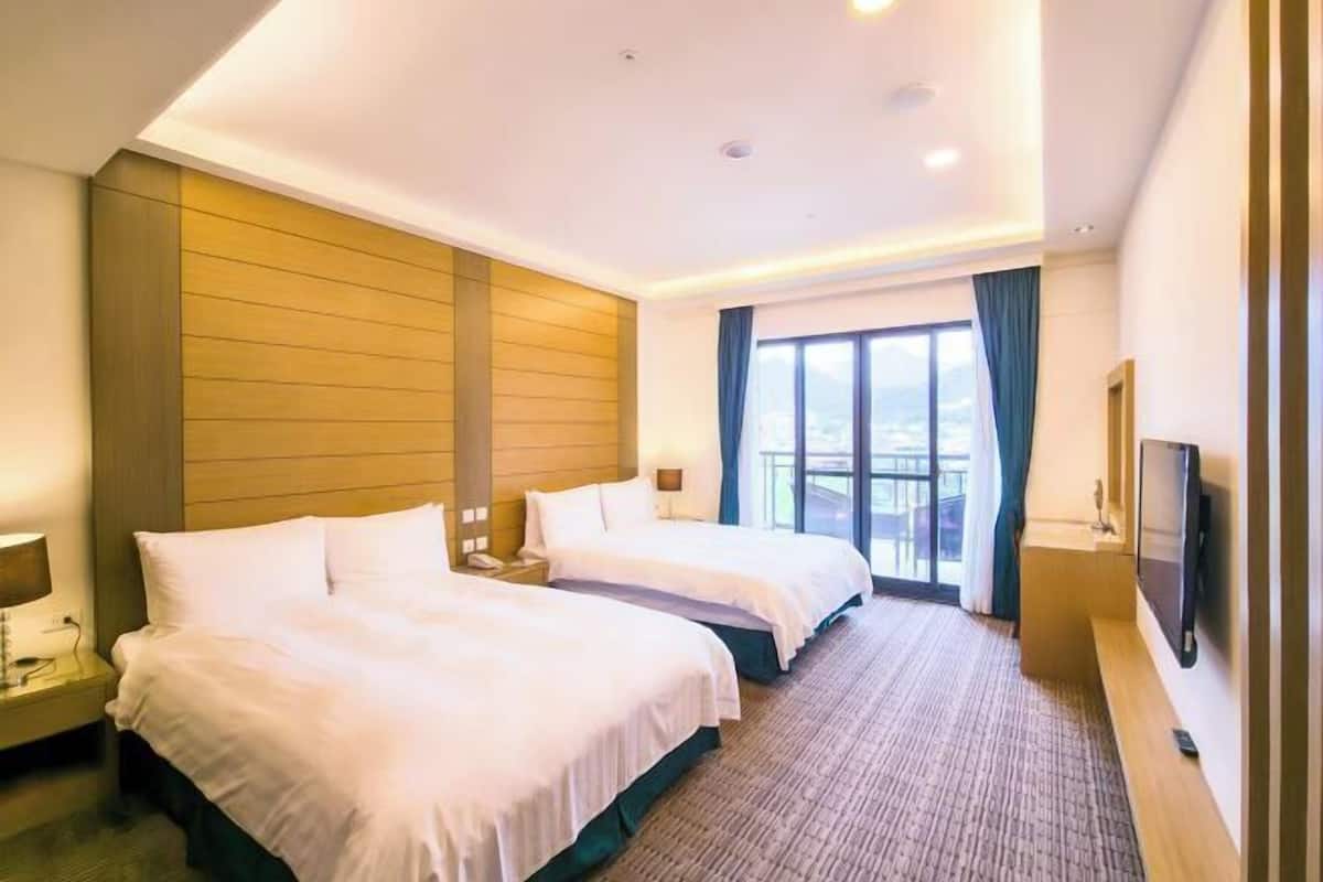 Habitación de hotel con dos camas, televisión, escritorio y balcón con vistas.