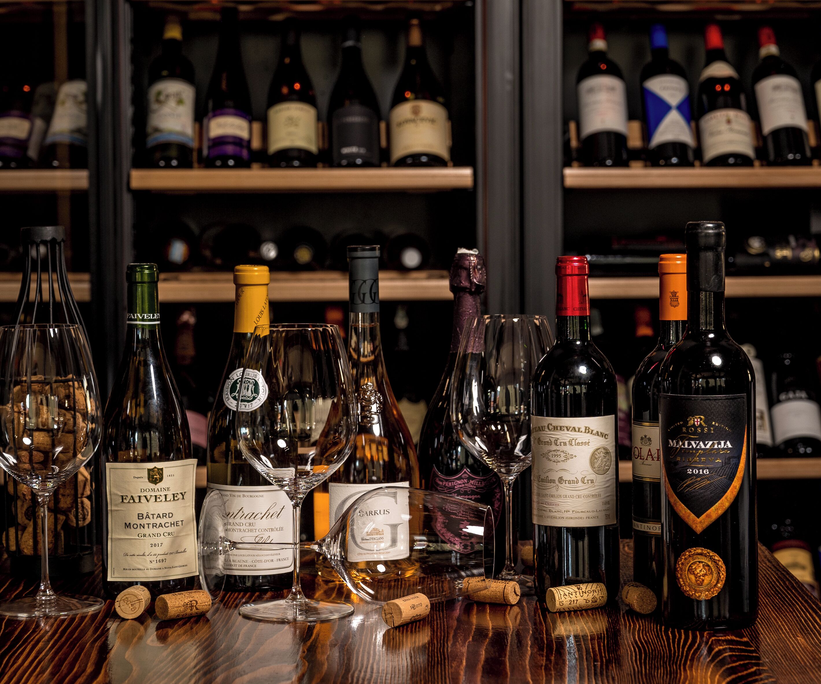 A selection of wine bottles and glasses on a wooden table, with shelves of wine bottles in the background.