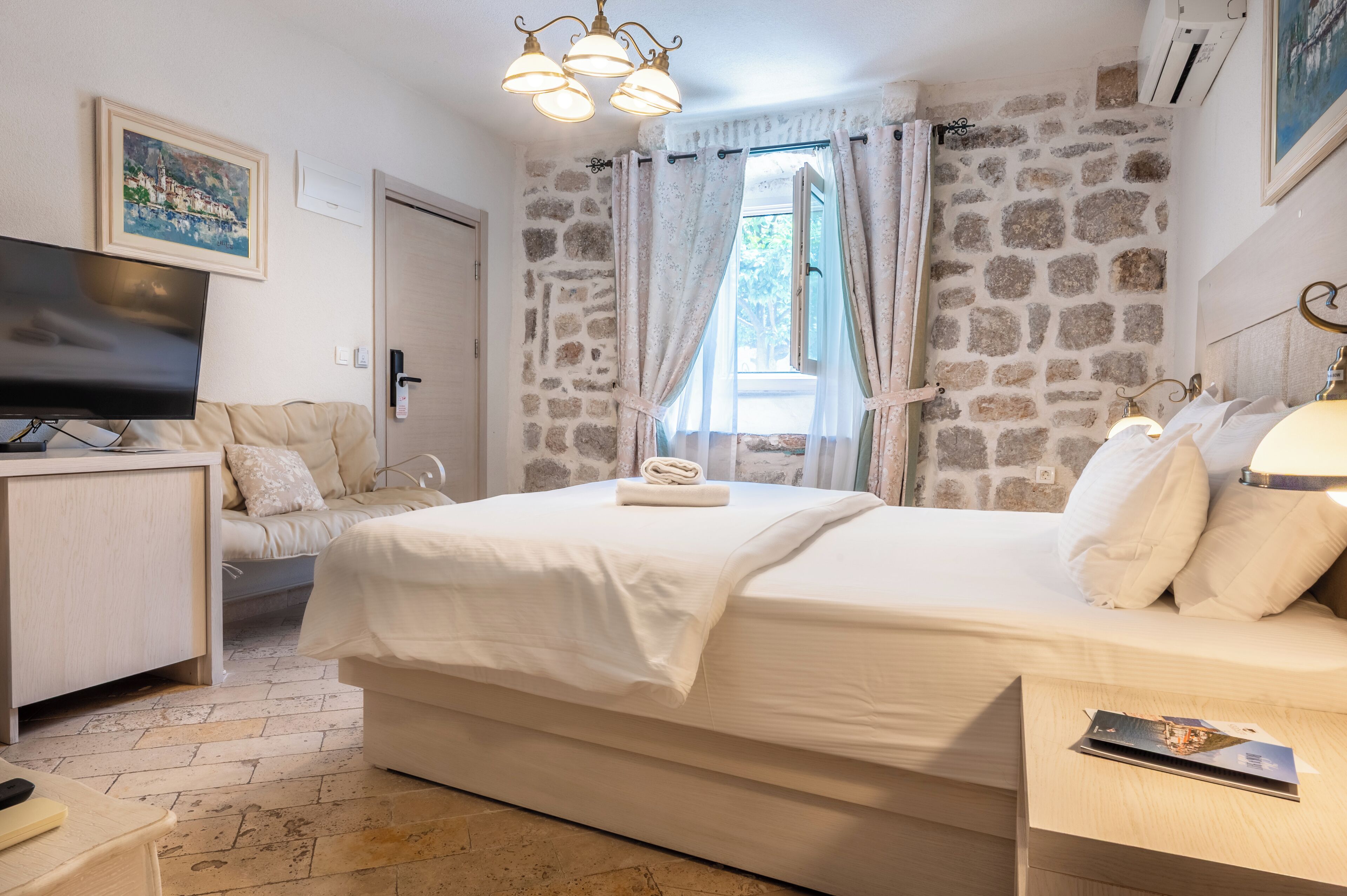 Un dormitorio con pared de piedra, cama con ropa de cama blanca, televisor en una mesita de noche y un cuadro en la pared.