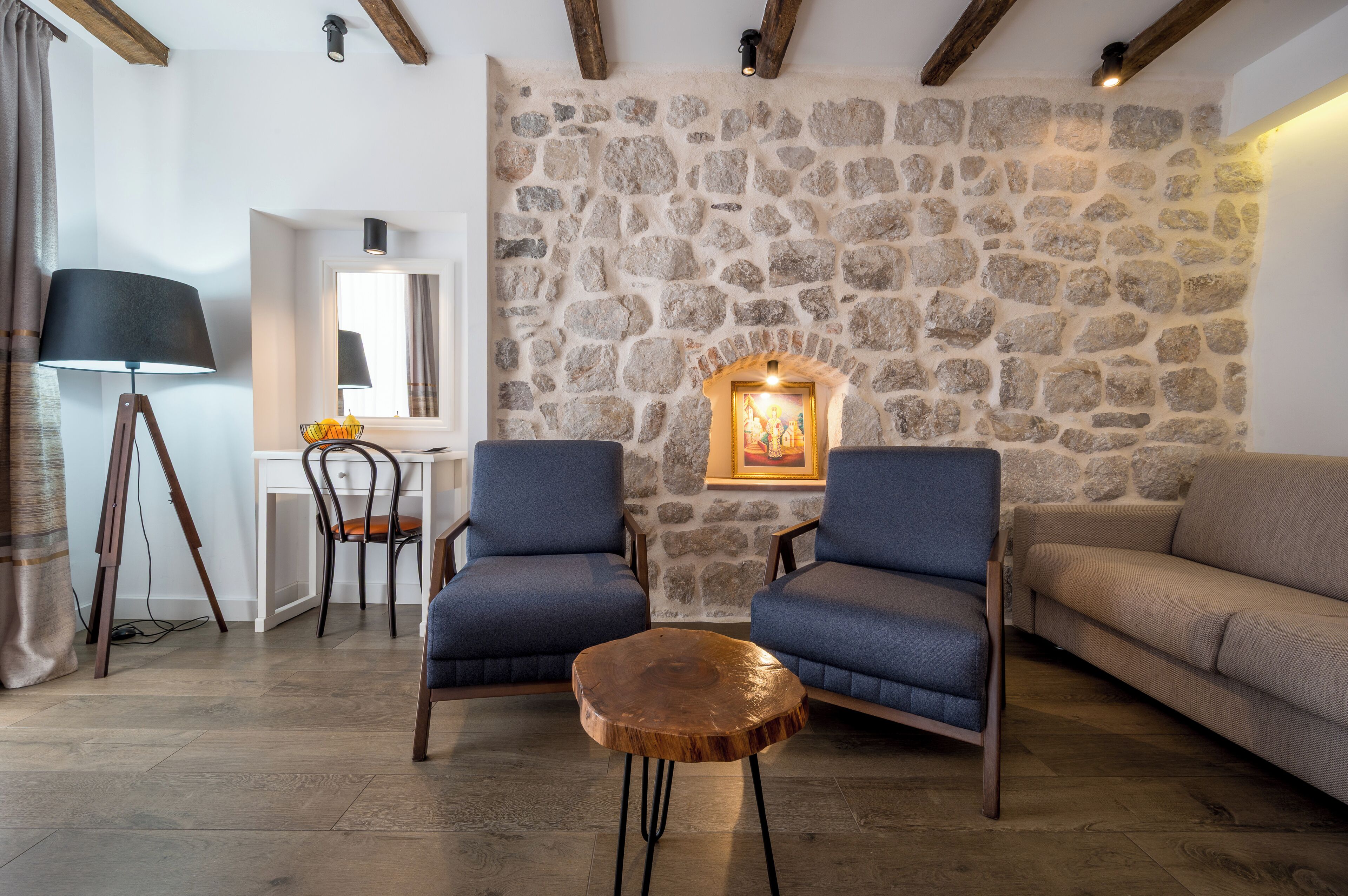 Una sala de estar acogedora con una pared de piedra, dos sillones azules, una mesa de centro de madera, un escritorio blanco y un sofá beige.