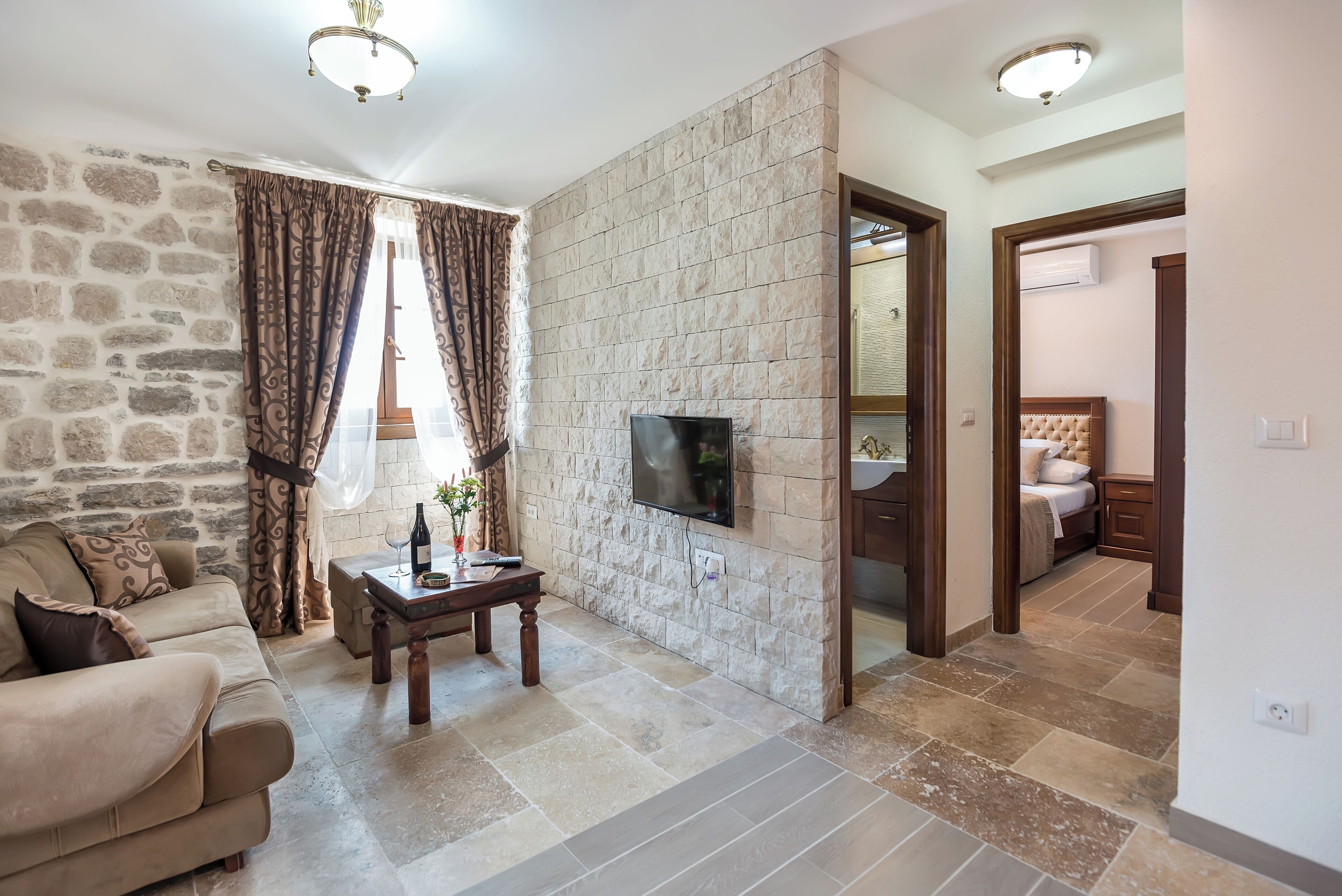A living room with a stone wall, a flat-screen TV, a sofa, a coffee table, and a window with curtains.